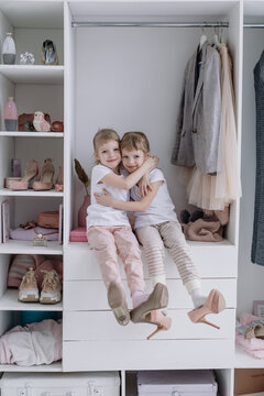 Family wardrobe. Happy twin sisters on shelve in closet with natural organic clothes in cozy dressing room play joyfully