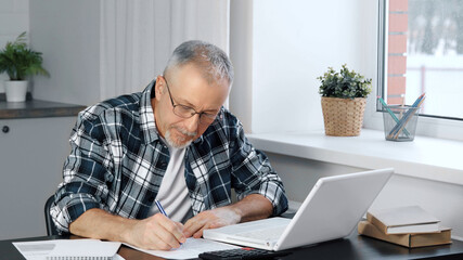 A thoughtful elderly man sits at his laptop, looking through documents.