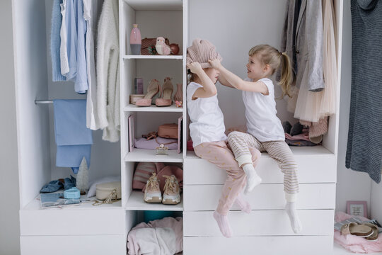 Family wardrobe. Happy twin sisters on shelve in closet with natural organic clothes in cozy dressing room play joyfully