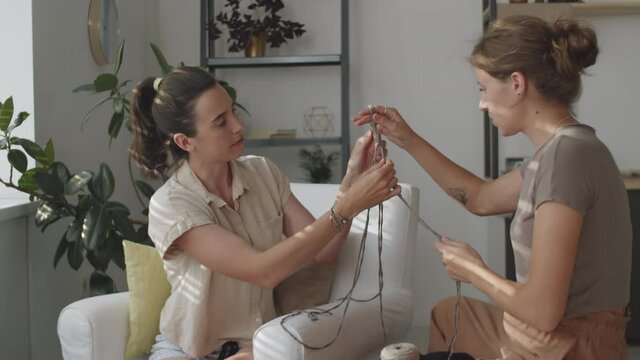 Two young Caucasian women knotting macrame plant hanger while spending day together at home