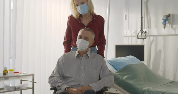 Senior Wife In Protective Mask Pushing Discharged Male Patient On Wheelchair In Hospital