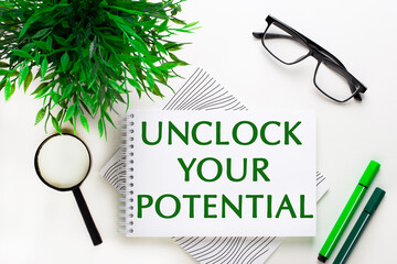 On a white background lies a notebook with words UNLOCK YOUR POTENTIAL, glasses, a magnifying glass, green markers and a green plant