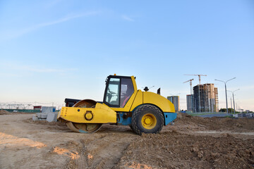 Fototapeta na wymiar Vibro Roller Soil Compactor leveling ground at construction site. Vibration single-cylinder road roller on construction road. Road work for new asphalt laying. Tower cranes build high-rise buildings