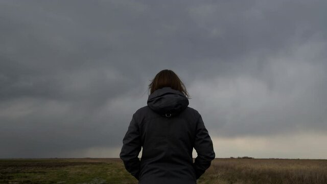 Woman standing in meadow, looking at the horizon and dark dramatic stormy clouds, rear view