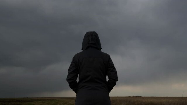 Rear view of female person wearing hooded jacket against dark moody dramatic clouds at sky