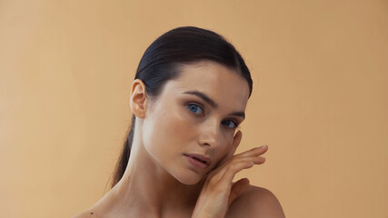 young woman with healthy skin touching face while looking at camera isolated on beige