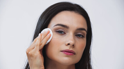 brunette woman wiping face with cotton pad while looking at camera isolated on grey