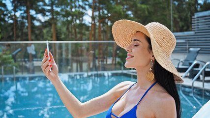 happy brunette woman in straw hat taking selfie near pool