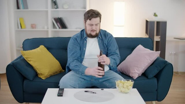 Obese man opening glass bottle of beer, drinking alcohol in front of tv at home