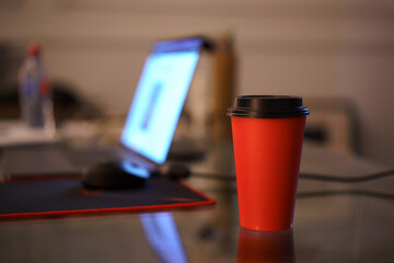 red paper cup in the office on the table in the evening with lamp light on the background of a glowing laptop. Work space and overtime.