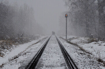 railway track in the forest, snowfall in spring, semaphore shows yellow light, people walk on the...