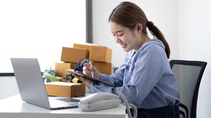 Young Asian woman is writing down the customer's details and addresses on the Clipboard in order to prepare for shipping according to the information, New kind of business for young, Sell online.
