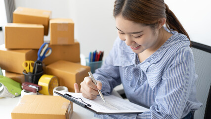 Young Asian woman is writing down the customer's details and addresses on the Clipboard in order to prepare for shipping according to the information, New kind of business for young, Sell online.