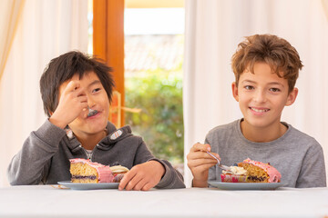 Two multi racial children eating cake, celebrating and laughing inside a house