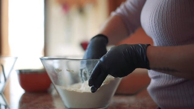 Detalle de una mujer con guantes negros moviendo la harina dentro de un recipiente de cristal transparente en la encimera de la cocina.