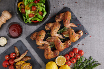 Roasted chicken wings in barbecue sauce and mixed vegetable salad with pepper seeds rosemary, salt in black stone plate on gray stone table. top view with copy space. tasty snack. Flat lay.