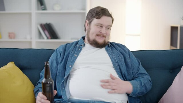 Lonely obese man relaxing on sofa at home, drinking alcohol, harmful addiction