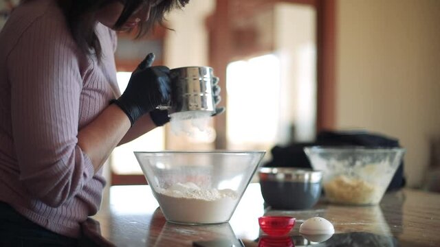 Una mujer con guantes en la cocina usando un utensilio para tamizar la harina que cae lentamente en un bol de cristal.