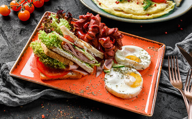 Delicious breakfast with fried eggs, sandwiches with ham and vegetables, and sausages served on plate over dark background. Healthy breakfast concept, close up