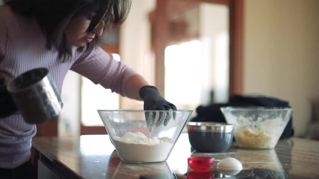Una mujer con guantes en la cocina usando un utensilio para tamizar la harina que cae lentamente en un bol de cristal.