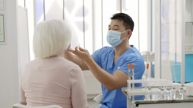 Male doctor in protective mask taking PCR test sample from throat of female retiree patient and putting it in glass tube and scanning. . Concept of Covid diagnostic.