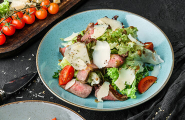 Tasty steak salad with tomatoes, green leaves, and hard cheese on dark background close up view. Concept for a tasty and healthy meal