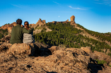 Young couple from behind sitting and watching Roque Nublo, symbolic natural monument of Gran Canaria. Summer sunny day blue skies and moon. Emblematic volcanic rock formations Canary Islands Spain.