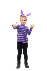 Cute little girl with Easter bunny ears looking up holding painted egg showing thumb up. Full body isolated on white background