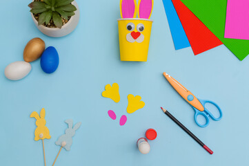 Happy Easter day.Festive craft from a paper cup with your own hands.Leisure for children.A congratulatory gift.Cute yellow Easter bunny on blue background