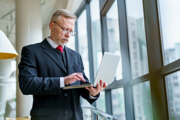 Fototapeta premium Thoughtful businessman standing in cafe with modern laptop. Aged man in dark suit red tie and glasses.