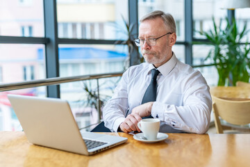 Fototapeta na wymiar Wise businessman siting in front of modern laptop, Adult man in white shirt and working in cafe with cup of coffee.