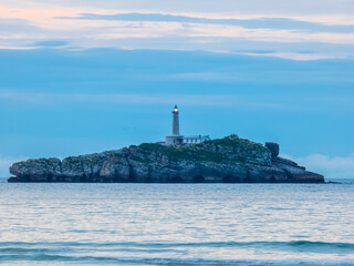 La luz del faro situado sobre una pequeña isla rocosa se observa desde la playa de Somo, España,...