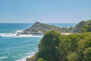 Fotobehang Grandes piedras en las costas e isla llena de árboles y palmeras mar azul claro con grandes olas blancas en las playas de Tayrona con la jungla a la izquierda © Esteban.94c