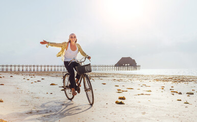 Young female dressed light summer clothes joyfully threw up her hand riding old vintage bicycle with front basket on the low tide ocean white sand coast on Kiwengwa beach on Zanzibar island, Tanzania.