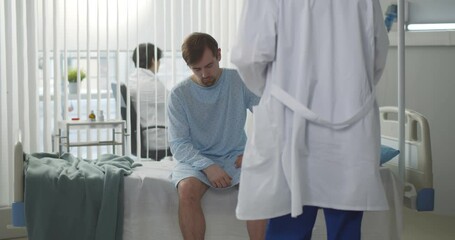 Sad young male patient signing medical form sitting on hospital bed in modern clinic.