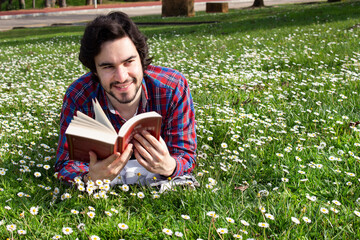 Smiled young man with unfocused book lying on the grass field with flowers around. Young man reading a book