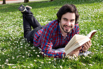 Smiled young man with unfocused book lying on the grass field with flowers around. Young man reading a book