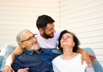 Happy senior couple sitting on sofa while his thankful adult brunette bearded son hugging them with love and care, enjoying weekend family time at home together. Generation and people concept