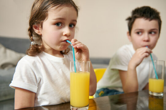 Adorable Children Drinking Orange Juice From Straw Sitting At The Table At Home