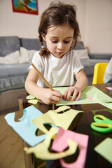 Preschool girl sitting at the table and concentrate on drawing shapes on colored paper at home. Children creativity concept
