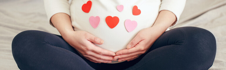 Closeup of pregnant woman in white clothes sitting on bed touching her belly. Pregnant belly covered with red pink paper hearts. Expecting young lady at home. Web banner header.