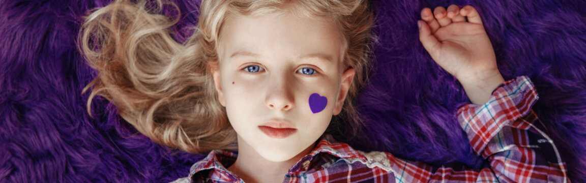  International world epilepsy illness awareness day. Cute pretty blonde Caucasian girl with small violet purple paper heart on her cheek lying on purple fluffy rug carpet at home. Web banner header.