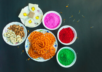 Colorful holi powder or gulal in ceramic bowl along with traditional Indian sweet food imarti, sandesh, cashew and almonds in plate on black background.