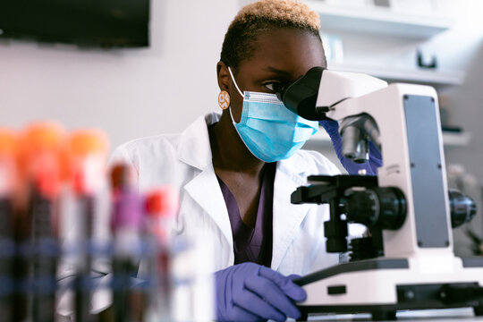 Black female scientist in mask in laboratory looking through microscope