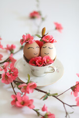 Creative easter eggs with cute face, sleepy eyes and spring flowers in tea cup on white background. Happy Easter concept, mockup, copy space. 