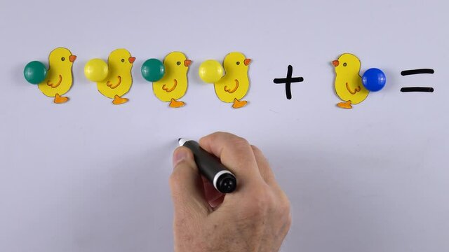 Teacher teaches kids basic of math addition with the aid of magnets and figures of chicks, cut out from paper, on the whiteboard, example of educational math children game