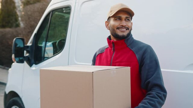 Delivery man smiling while holding parcel in front of transportation van. High quality 4k footage
