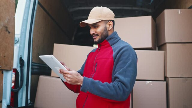 Young courier using tablet in front of the van full with cardboard boxes. Delivering the pardel. High quality 4k footage