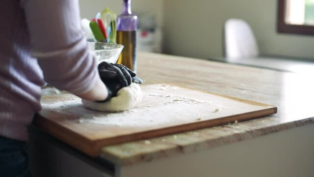 Una mujer en la cocina amasando una masa de pizza en una tabla de madera con harina mientras usa guantes negros.