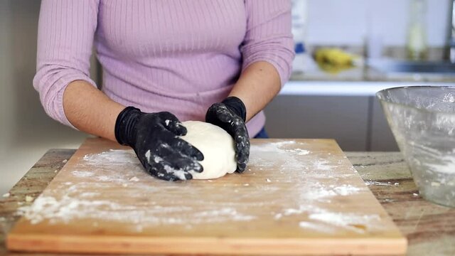 Una mujer en la cocina amasando una masa de pizza en una tabla de madera con harina mientras usa guantes negros.